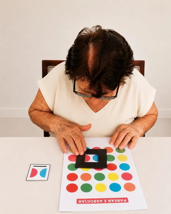 Senhora usando uma moldura de papel para encontrar, numa folha com círculos coloridos, o par igual ao cartão de referência.